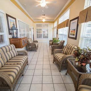 A sunlit interior space featuring wicker furniture and tiled flooring, with large windows and decorative elements.