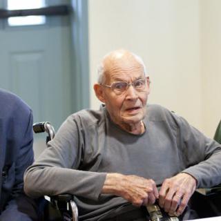 An elderly man wearing glasses is seated in a wheelchair indoors.