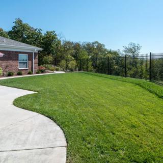 A well-maintained yard with a green lawn and a nearby house, enclosed by a fence under a clear, sunny sky.