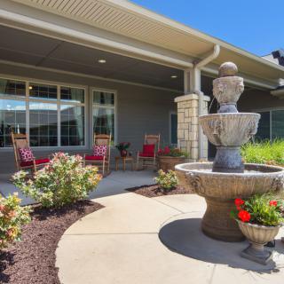 A sunny patio with a decorative fountain, surrounded by flowers and comfortable seating.