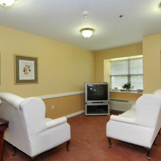 A cozy living room with two white armchairs facing a television, complemented by artwork on the walls and natural light from a window.
