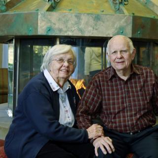 An elderly couple sitting together in front of a fireplace.
