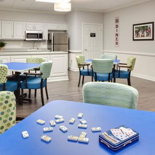 A modern dining and game room with tables, chairs, and a kitchen area featuring dominoes on one table.