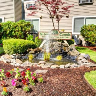 A charming garden area with a small fountain surrounded by flowering plants and greenery, located in front of a building.
