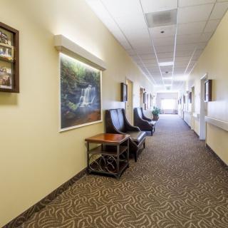 A hallway with chairs and tables along the wall, featuring carpeted flooring and artwork on the walls.