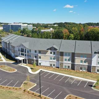 An aerial view of a modern residential apartment complex with an adjacent parking lot, surrounded by trees and other buildings in the background.
