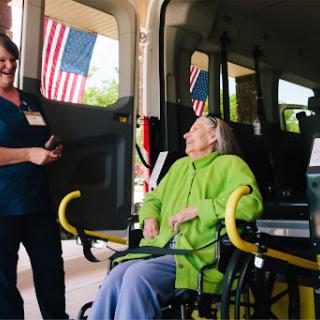 A caregiver interacts with a senior citizen in a wheelchair, preparing for transport outside a vehicle adorned with American flags.