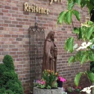 A statue stands against a brick wall surrounded by flowers and greenery.