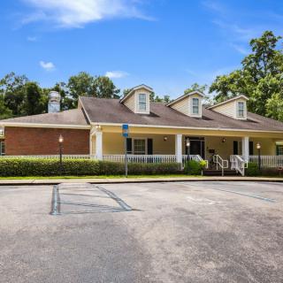 A single-story house with a parking lot in front, surrounded by trees under a blue sky.