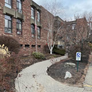 A winding pathway leads to a brick building, flanked by landscaped areas and a parking sign.