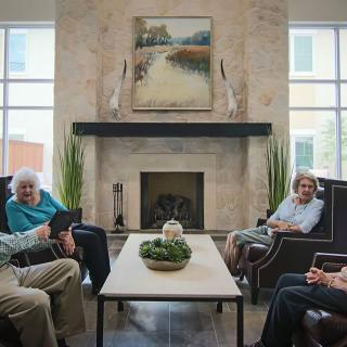 A group of seniors sitting in a cozy living room, engaged in conversation near a fireplace.