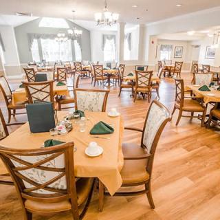 An elegantly set dining room with multiple tables and chairs, featuring a warm wooden floor and natural lighting.