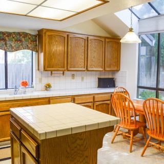 A bright kitchen with wooden cabinets, a tiled island, and orange dining chairs under ample natural light.