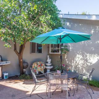 A cozy outdoor patio with a table and chairs under a large umbrella, surrounded by greenery and a decorative fountain.