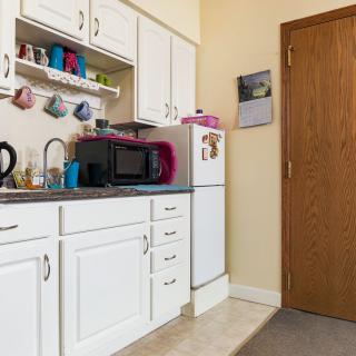 A compact kitchen with white cabinets, a microwave, refrigerator, and assorted mugs hanging above the counter.