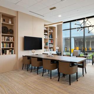 A modern conference room with a large table, comfortable chairs, and bookshelves, featuring a television and large windows for natural light.