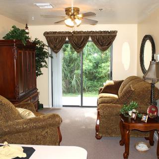 A cozy living room featuring a ceiling fan, plush sofas, and a large window with curtains, overlooking greenery.