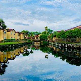A serene view of buildings and trees reflected in a calm river under a blue sky.