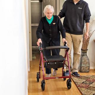 An elderly woman using a walker, supported by her caregiver as they move through a hallway.