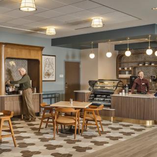 A cozy cafe interior with two people behind the counter and a seating area with tables and chairs.
