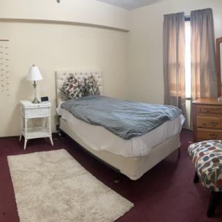 A cozy bedroom with a single bed, nightstand, and lamp, featuring warm curtains and carpeting.