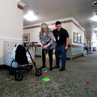 An elderly woman participates in a physical therapy activity with the assistance of a caregiver in a hallway setting.