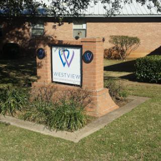 A brick sign displaying the logo and name of a building, surrounded by green landscaping.