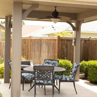 A cozy patio area with a table and chairs under a covered structure, surrounded by bushes and enclosed by a wooden fence.