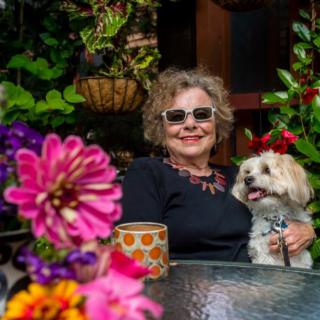 A woman sitting at a table with her dog, surrounded by vibrant flowers in a garden.