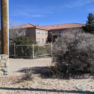 A view of a residential building surrounded by desert landscape, enclosed by a fence under a clear blue sky.