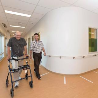 An elderly person using a walker is accompanied by a healthcare professional in a rehabilitation facility.