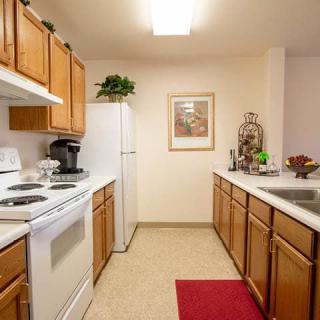 A cozy kitchen with wooden cabinets and white appliances, featuring a red rug and decorative elements.