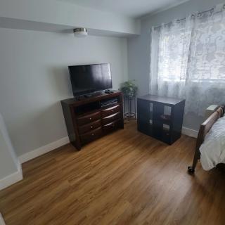 A cozy bedroom with a TV on a dresser, hardwood floors, and curtains by the window.