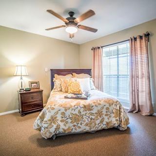 A cozy bedroom with a bed, side table, and ceiling fan, featuring natural light from a large window with curtains.