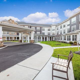 A modern three-story building with a circular driveway and an outdoor bench under a partly cloudy sky.