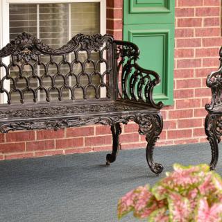 A decorative wrought iron bench and chair on a brick patio with green shutters.
