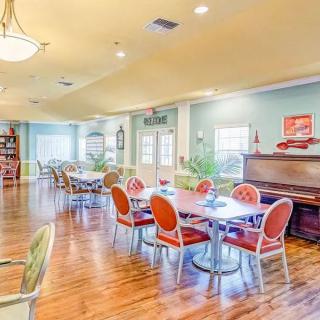 A well-lit dining area with colorful chairs, tables, and a piano against hardwood flooring.