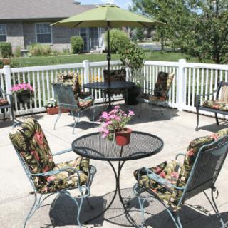 A cozy patio with colorful chairs and an umbrella, surrounded by a white fence and potted plants.