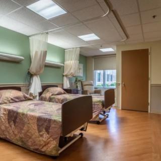 A hospital room with two beds separated by curtains, featuring a wooden floor and bright lighting.