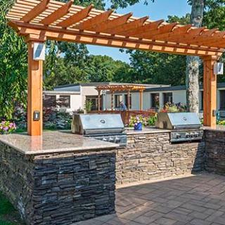 An outdoor kitchen setup with a pergola, featuring two barbecue grills and a stone countertop on a patio.