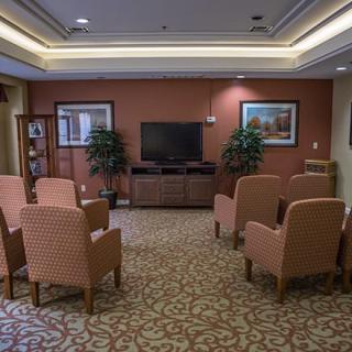A cozy lounge area with arranged chairs facing a television, featuring decorative elements like plants and paintings.