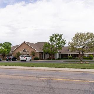 A suburban building with a parking lot, surrounded by trees and cars on a cloudy day.