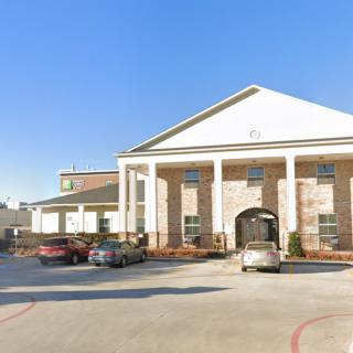 A large building with columns and a parking lot in front, featuring several parked cars on a clear day.