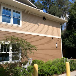 A two-story building with siding exterior, surrounded by greenery and trees on a sunny day.