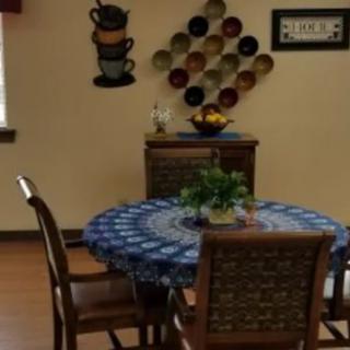 A cozy dining area with a round table covered by a blue patterned tablecloth, surrounded by chairs. The walls are decorated with artistic cup designs and a collection of colorful plates. A fruit bowl and a small indoor plant add to the homely atmosphere.