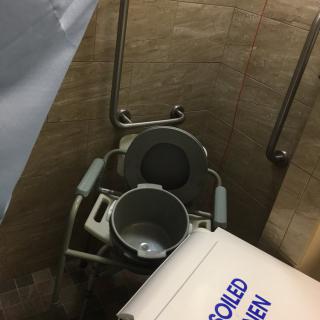 A bathroom equipped with a commode chair, bucket, and cooler.