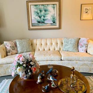 A cozy living room features a plush cream sofa adorned with patterned cushions, a wooden coffee table displaying a floral centerpiece, and elegant wall art.