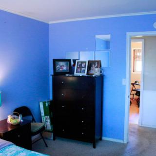 A cozy bedroom with blue walls, featuring a dark dresser adorned with photographs and a chair beside it.
