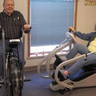 Two elderly individuals exercising indoors on stationary bikes.