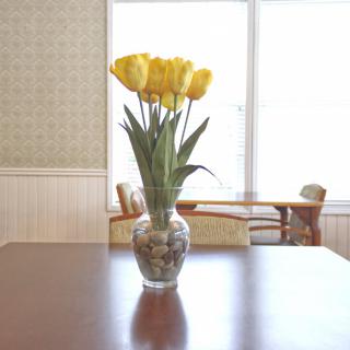 A bouquet of yellow tulips in a decorative vase sits on a wooden table in a well-lit room.